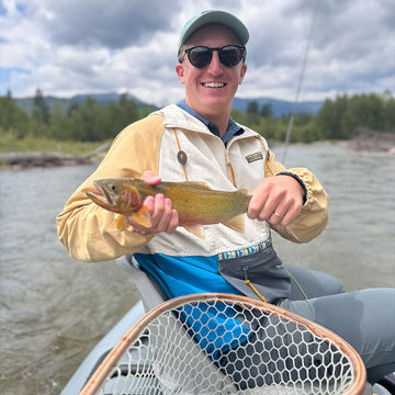 Man holding a fish caught while fishing on a river with mountains in the background